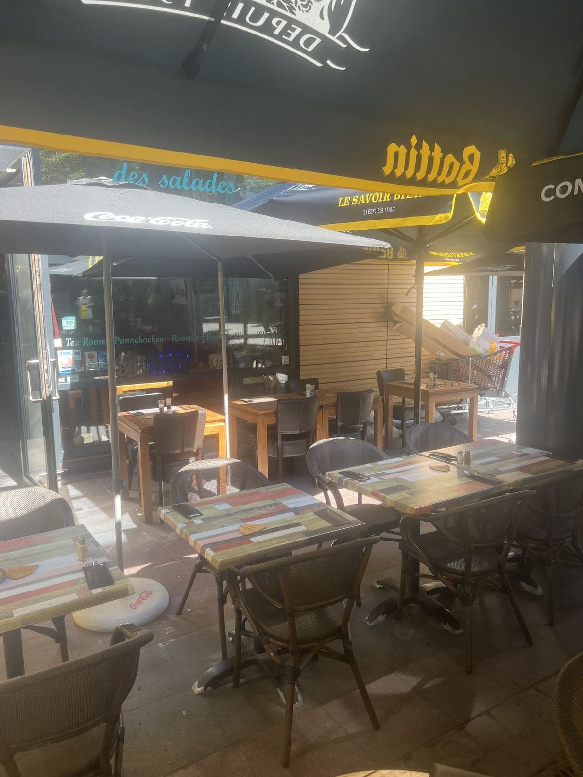 La terrasse abritée devant la vitrine où l'on lit « des salades », tables en bois peint et chaises tressées