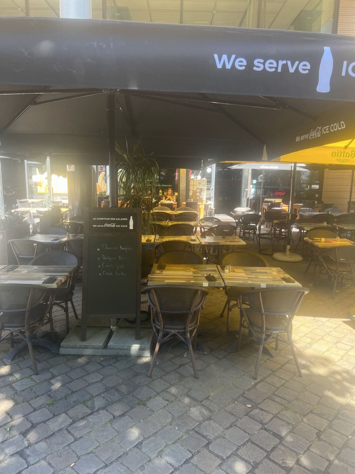 Het terras in de volle zon onder grote zwarte parasols, de gedekte tafels op de kasseien van het plein, het krijtbord van het restaurant bij de ingang