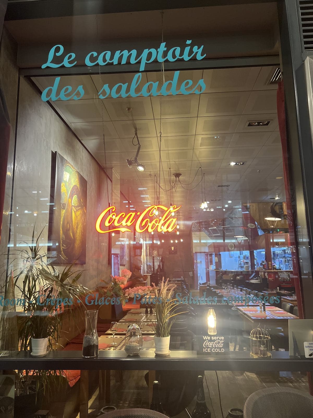 La vitrine du restaurant le soir : « Le comptoir des salades » en néon turquoise, une enseigne Coca-Cola allumée, la salle et ses tables derrière la vitre