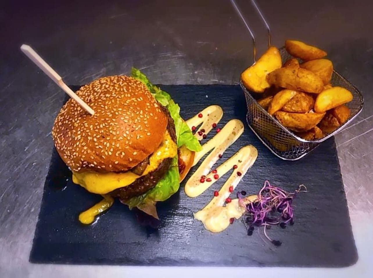 Un cheeseburger au pain au sésame sur une ardoise, un panier de pommes de terre rôties à côté, deux traits de sauce