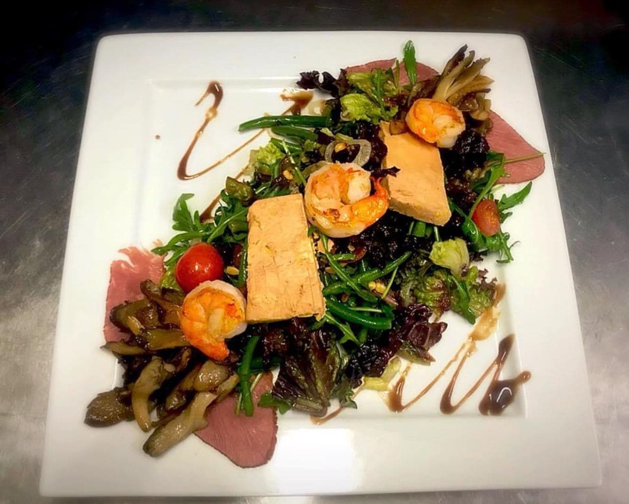 Een salade op een vierkant bord: foie gras, scampi's, gebakken champignons en eendenborst, onder strepen balsamicosaus