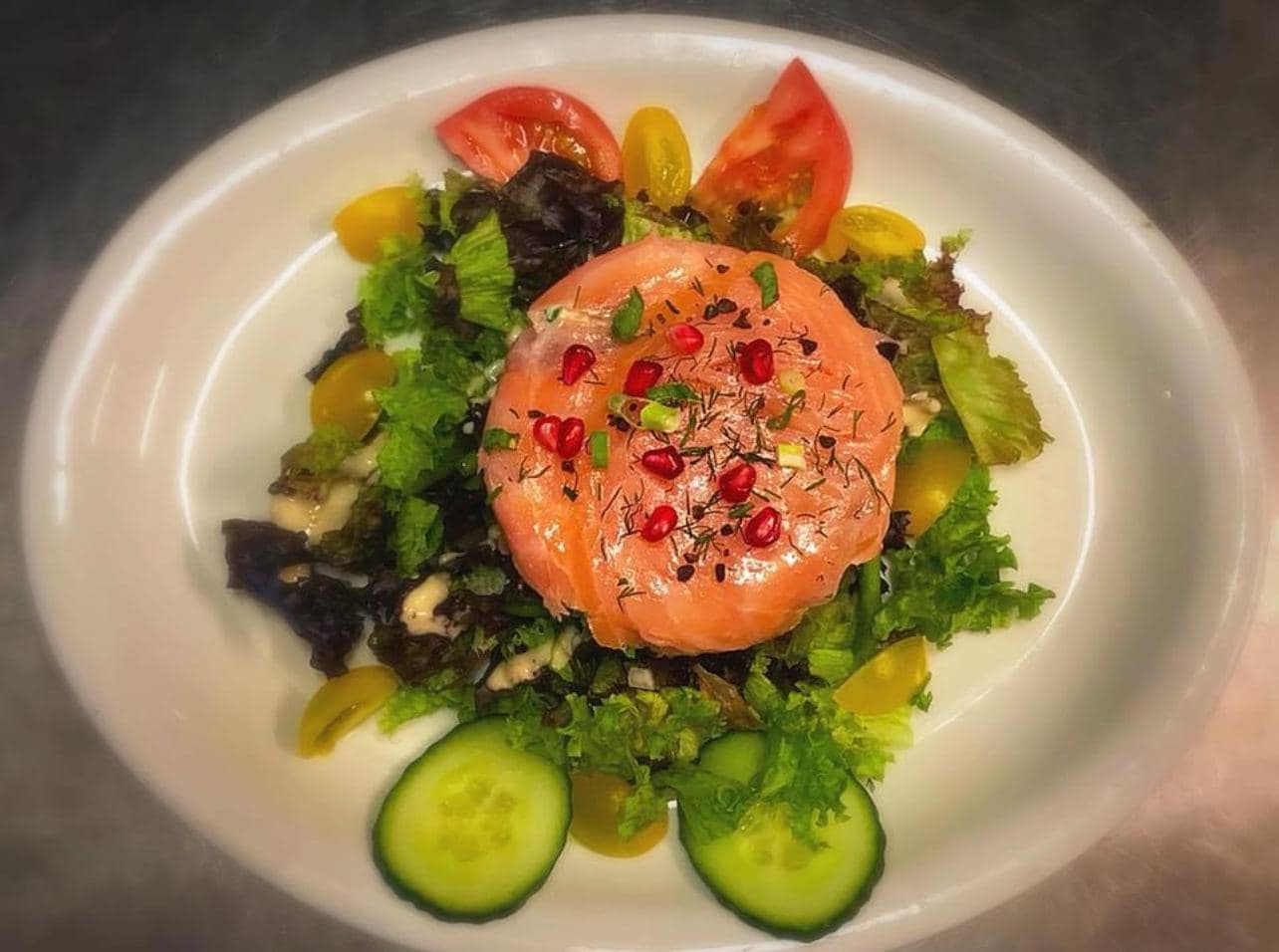 Een zalmtartaar met granaatappelpitjes, midden in een groene salade met tomaten en schijfjes komkommer