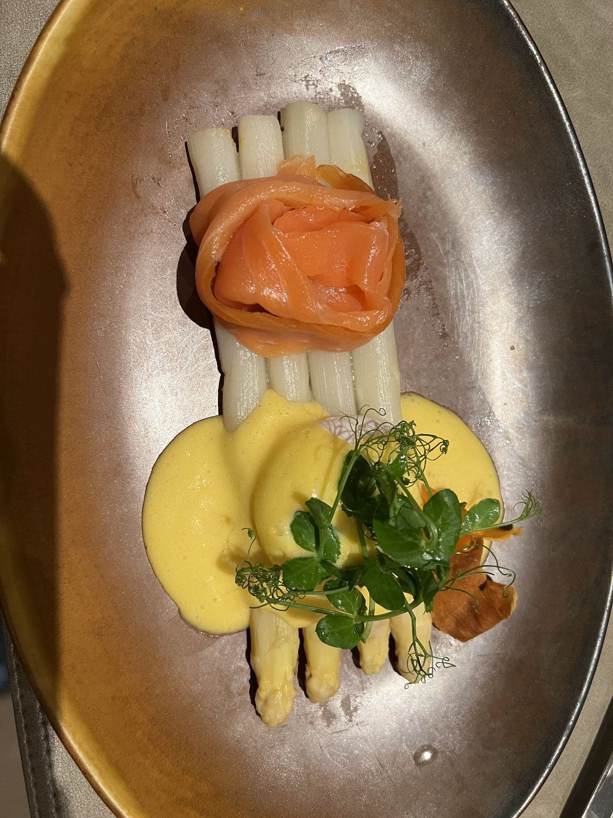 Witte asperges, een roosje gerookte zalm en een gele saus, op een ovaal koperkleurig bord