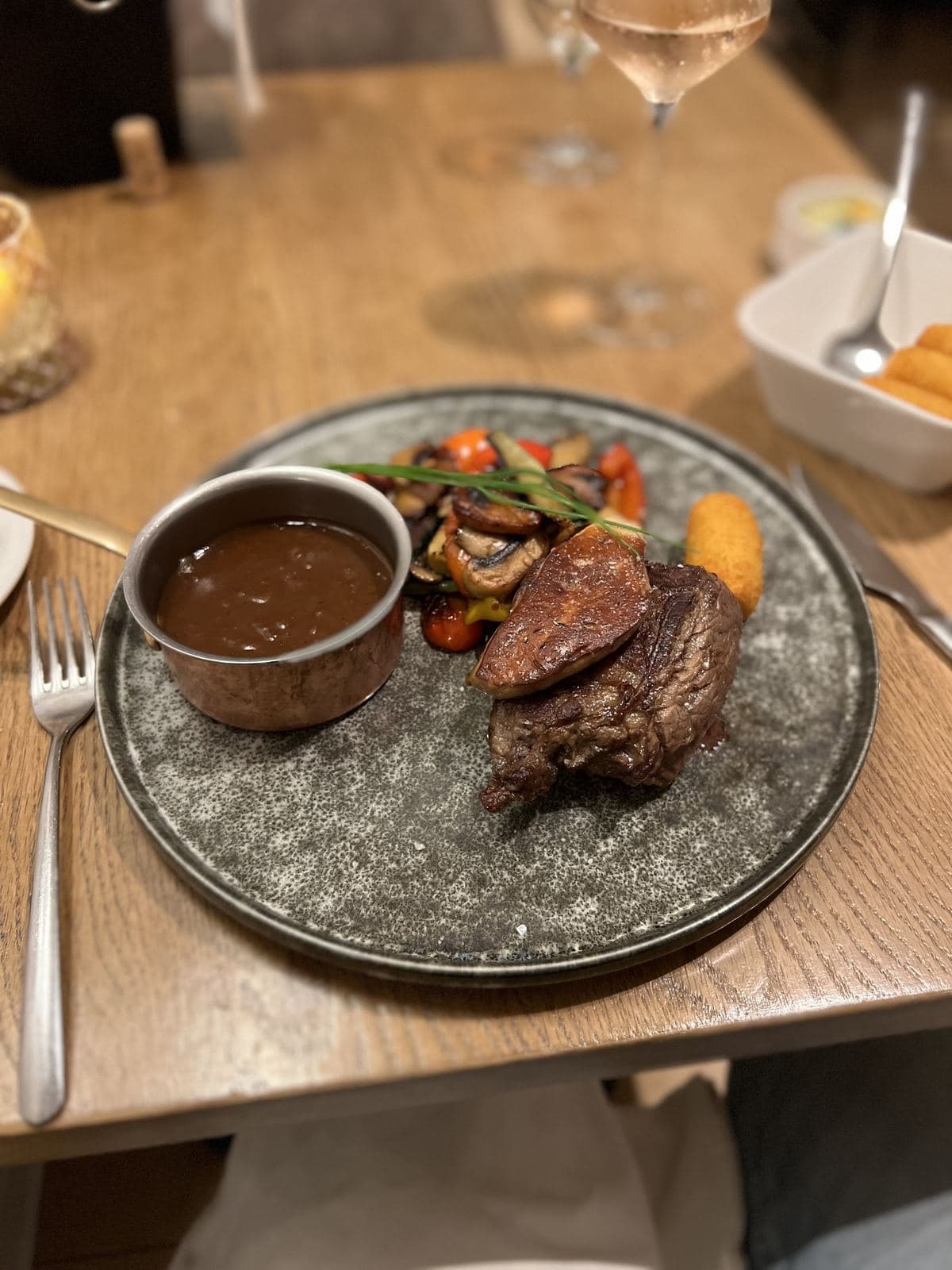 Une pièce de bœuf grillée, des légumes poêlés et une croquette, une saucière de sauce brune à côté, sur une table en bois