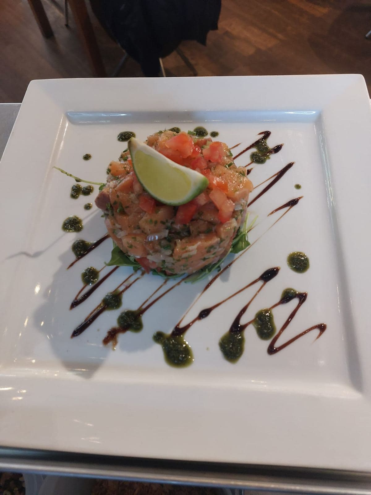 Een zalmtartaar met een schijfje limoen, op een vierkant bord met strepen saus